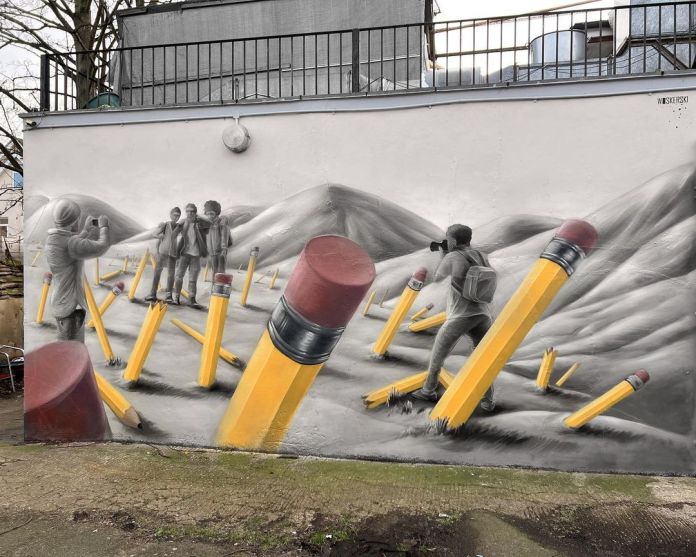 Street art mural in London showing giant yellow pencils planted in a grayscale mountain landscape with black-and-white figures photographing each other among the pencils.