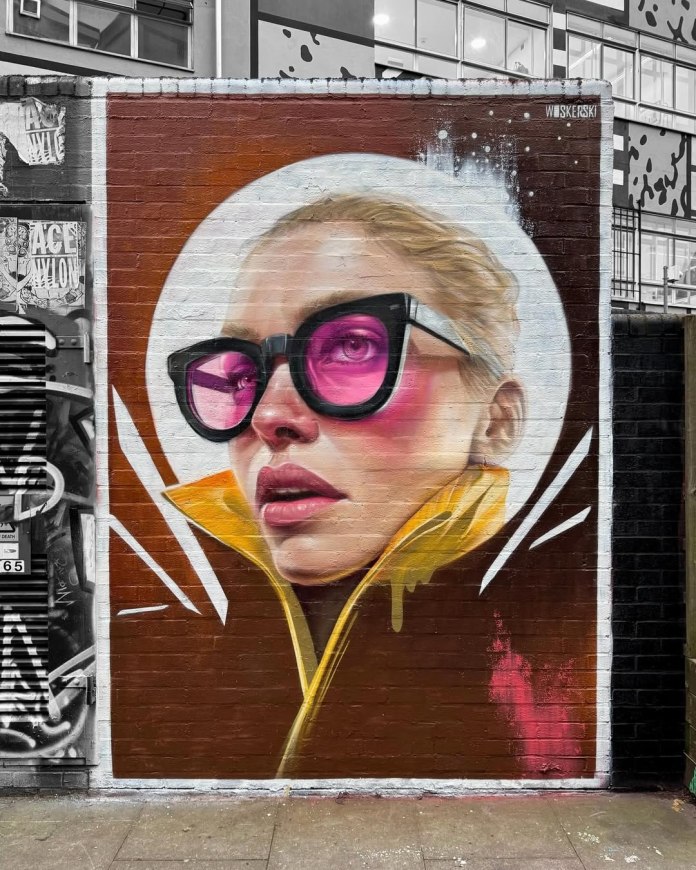 Mural of a stylized woman in angular black glasses with pink lenses, bright blush, and a yellow collar, set against a brown background with white accents.