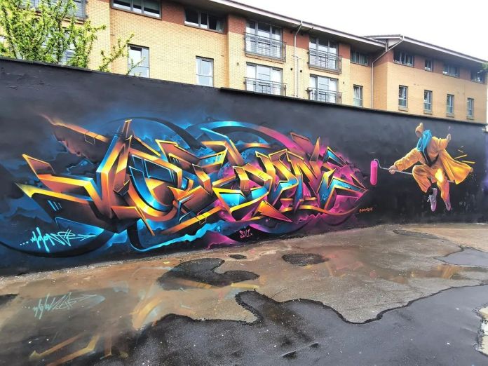 Wide shot of a collaborative mural in Glasgow with vibrant 3D graffiti text in yellow, blue, and pink tones next to a dynamic leaping figure in a yellow coat.