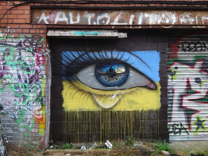 A close-up mural by My Dog Sighs in Cardiff depicting a detailed blue and yellow Ukrainian-flag-colored eye. Reflected in the pupil is the city of Kyiv engulfed in flames. A painted tear drips down the cheek, symbolizing grief over the conflict in Ukraine.