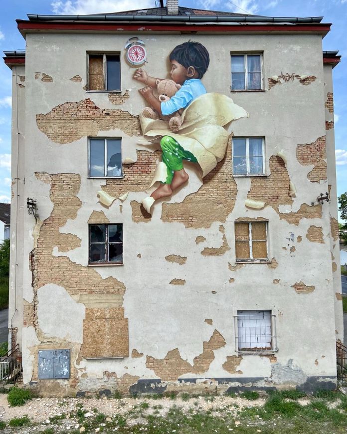 A realistic mural of a young boy holding a teddy bear, painted to appear as if peeling out of a building’s wall, in Plzeň, Czech Republic. The artwork integrates seamlessly with the aged facade, incorporating the building’s decay artistically.