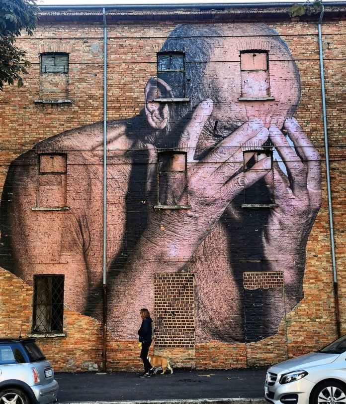 A detailed mural by Bifido depicting an elderly man covering his face with both hands, conveying despair. Painted on a brick building in Gambettola, Italy, the windows blend seamlessly into the artwork, accentuating the man's anguish.