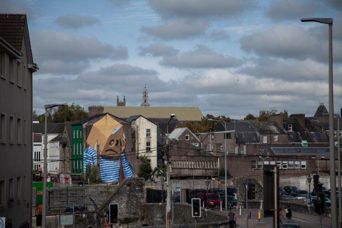 A mural by Asbestos in Cork, Ireland, portraying a figure wearing a cardboard box over their head. The box has eye holes revealing the person’s gaze, symbolizing anonymity and vulnerability in the context of Ireland's housing crisis.