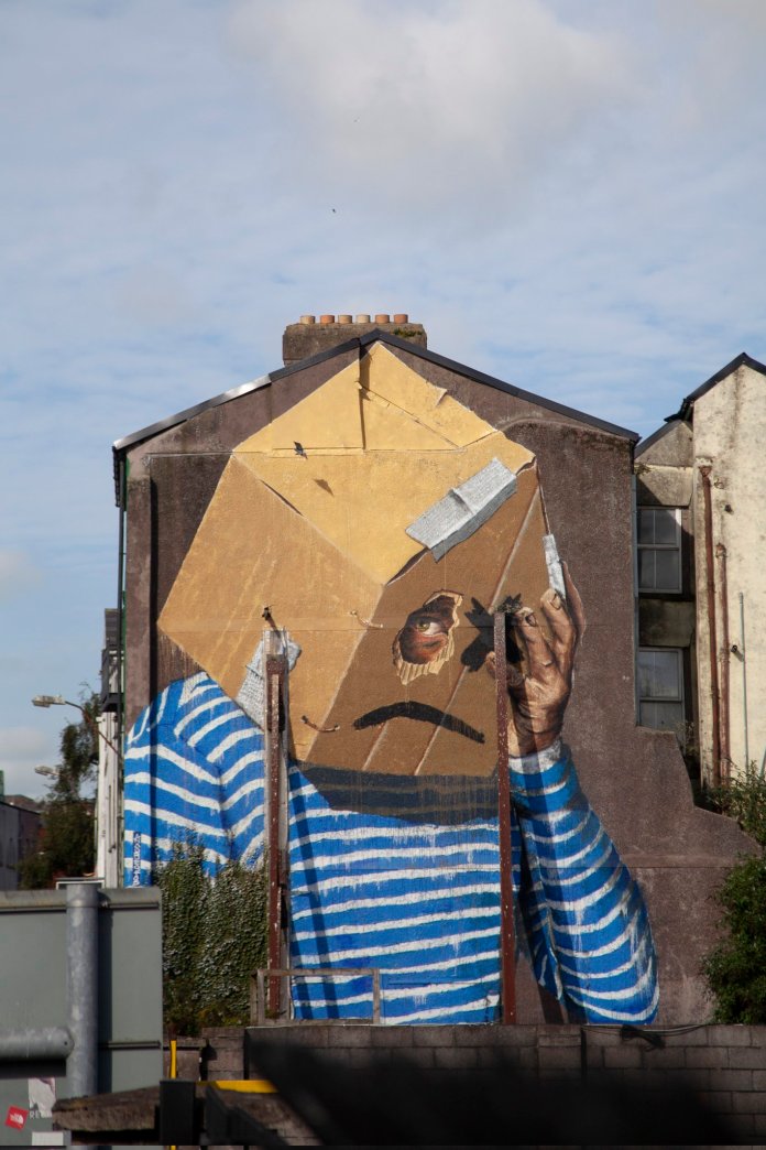 A mural by Asbestos in Cork, Ireland, portraying a figure wearing a cardboard box over their head. The box has eye holes revealing the person’s gaze, symbolizing anonymity and vulnerability in the context of Ireland's housing crisis.