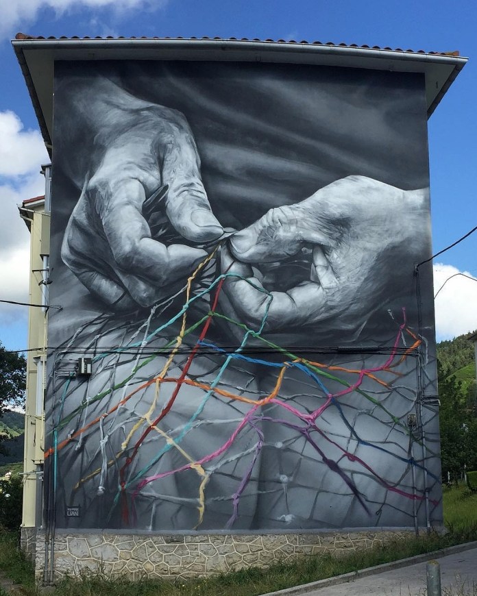 A grayscale mural of two elderly hands weaving a fishing net, painted by Murales Lian in Ondarroa, Spain. The hands are photorealistic, while the net is vividly colorful with strands in red, green, yellow, and pink, symbolizing tradition and community.