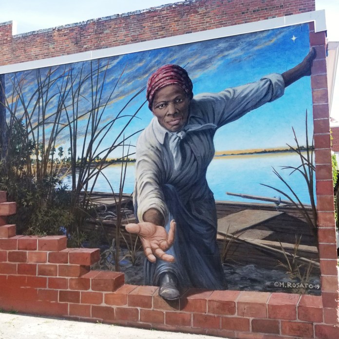 A realistic mural of Harriet Tubman painted by Michael Rosato in Cambridge, Maryland. She extends her hand through a painted break in a brick wall, set against a riverside landscape at sunset. The mural is titled Take My Hand and pays tribute to her legacy.