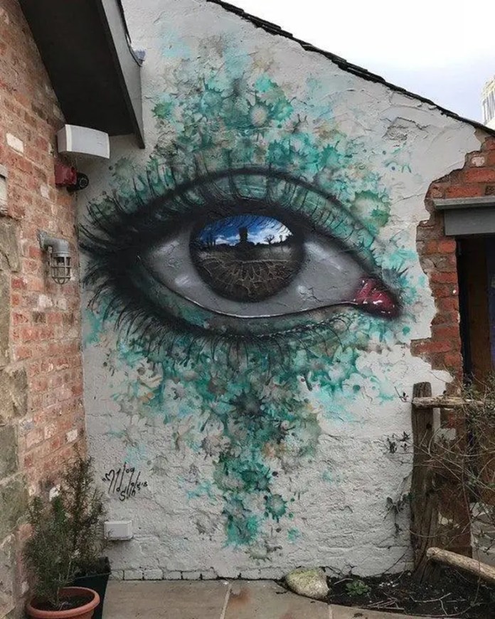 A street mural by My Dog Sighs in Eccleston, Lancashire, showing a detailed human eye with teal tears running down a rough, cracked wall. The reflection in the pupil captures a cobblestone path and a lone figure, creating a powerful sense of isolation and reflection.