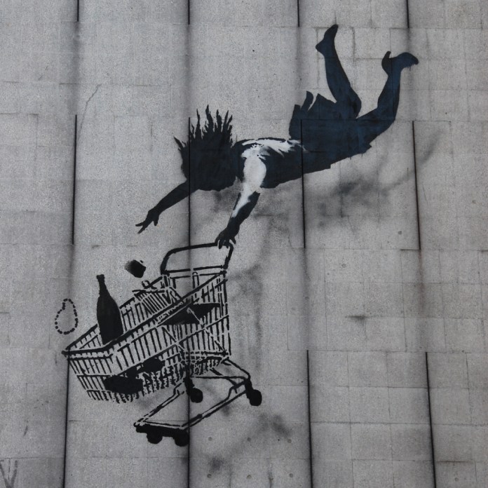 Banksy’s artwork Shop Until You Drop features a stencil of a woman falling headfirst through the air, clinging to a shopping cart filled with groceries. The piece critiques consumerism, portraying the dangerous obsession with material goods.