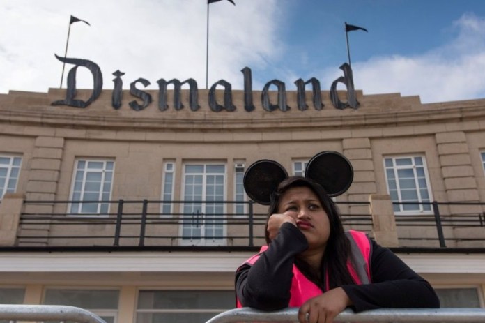A photograph of a staff member at Banksy’s Dismaland installation in 2015, wearing Mickey Mouse-style ears and a bored expression while leaning on a barrier. The background shows the dystopian theme park entrance with "Dismaland" in gothic lettering, critiquing the superficiality of commercial entertainment.