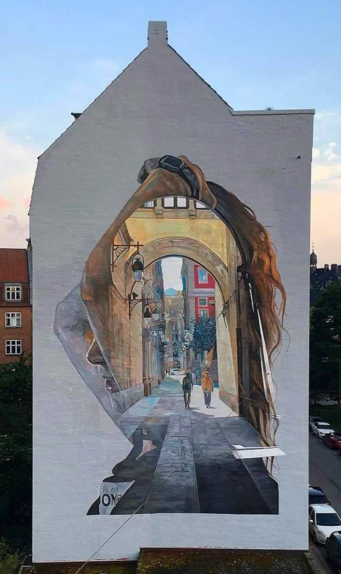 Large mural by Cristian Blanxer painted on the side of a building in Aarhus, Denmark, for the 17 Walls project. The artwork combines realism with surrealism, depicting the silhouette of a woman looking upwards. Inside her profile is a beautifully rendered street scene, as if viewing the scene through her mind’s eye. The detailed scene within the silhouette shows a European street with tall, historic buildings, arched architecture, and two pedestrians walking down a cobblestone path. The soft, warm lighting adds a sense of depth and tranquility to the piece, blending the woman's introspective expression with the urban landscape.