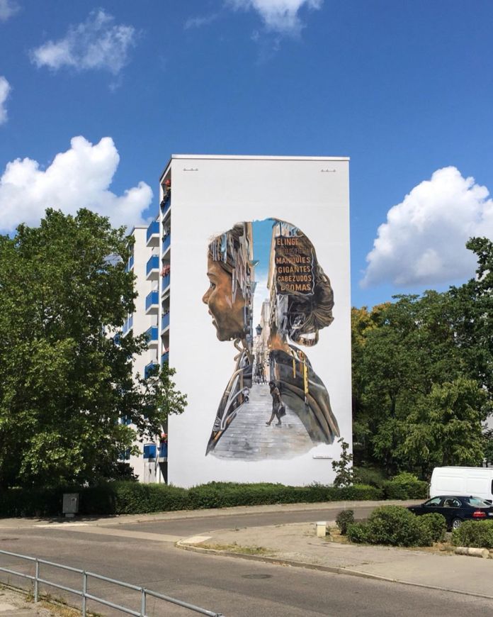 Mural titled 'Little Giants' by artists Cristian Blanxer and Victor García Repo on a building wall in Berlin, Germany, curated by Yasha Young. The artwork features a woman's profile, intricately filled with an urban street scene as if seen within her silhouette. This scene includes a pathway with pedestrians walking down steps, surrounded by signs and the architecture of a bustling city. The piece symbolizes a blend of introspection and urban life, reflecting on the modern human experience of being lost in everyday decisions and seeking deeper meaning. Cristian Blanxer's inspiration behind the mural evokes the idea of reconnecting with simple, natural joys—like noticing the blue sky above—as a reminder to find balance and perspective amidst the complexities of city life.