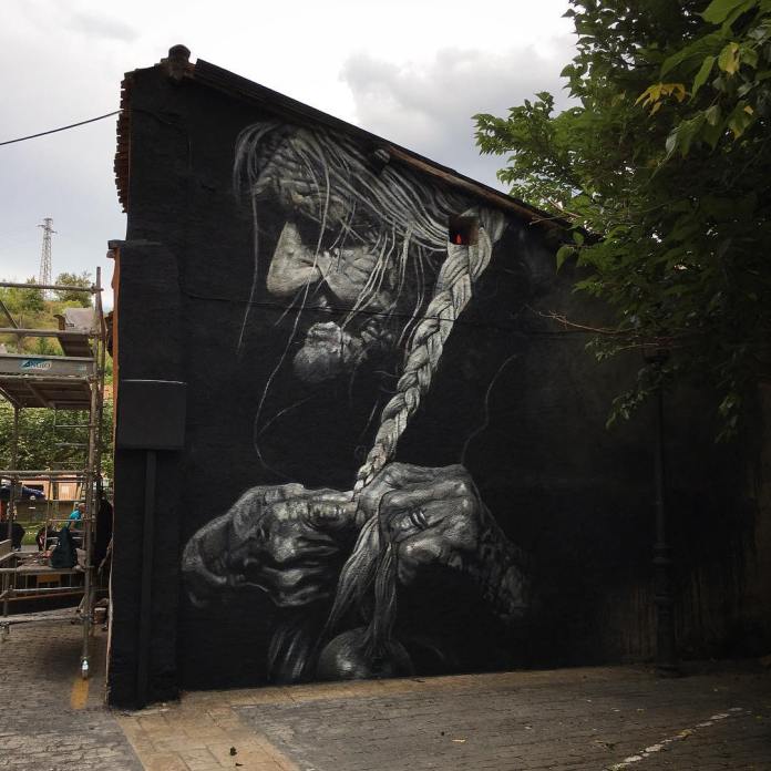 Mural titled 'Tipularia' (La cebollera) by artists Cristian Blanxer and Victor García Repo, created for the Cantamañanas Festival in Huarte, Navarre, Spain. The artwork, painted in grayscale, depicts an elderly woman with deep wrinkles and weathered hands, focused on braiding her hair with a sense of patience and quiet dignity. The mural captures intricate details in her hands and face, highlighting textures and shadows that convey the passage of time and life experience. The mural's dark background and realistic shading make it appear almost like a photograph against the building wall, blending beautifully with the surrounding natural environment.