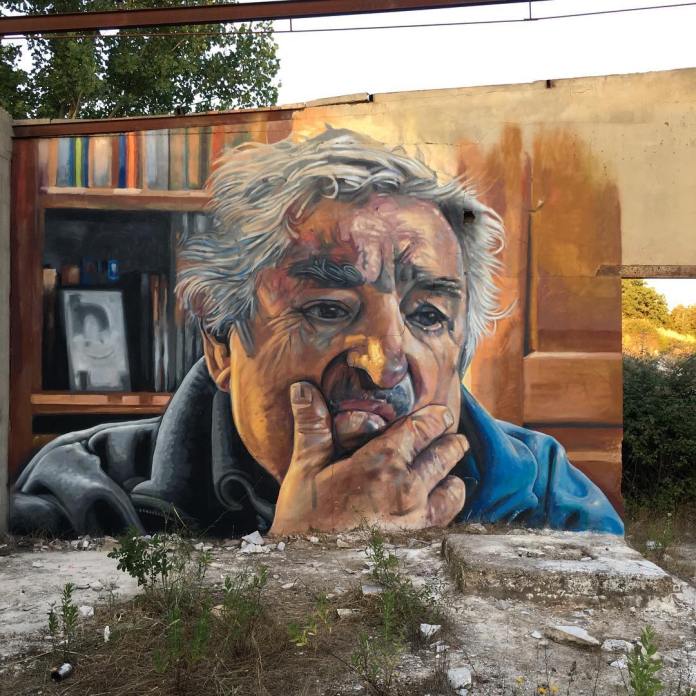 Portrait mural titled 'Mujica Maestro' by artists Cristian Blanxer and Victor García Repo, depicting José 'Pepe' Mujica, former president of Uruguay, located in Aracena, Spain. The detailed artwork shows Mujica deep in thought, with his hand resting thoughtfully on his chin, highlighting the lines and character in his face. In the background, a bookcase and warm earthy tones add context to the contemplative scene. A quote from Mujica is associated with the mural: 'No soy pobre, soy sobrio, liviano de equipaje, vivo con lo justo para que las cosas no me roben la libertad' (I am not poor, I am sober, light on baggage, I live with just enough so that things do not steal my freedom).