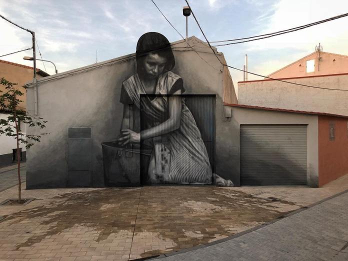 Street mural titled 'Más vale maña…' by artists Cristian Blanxer and Victor García Repo, located in Alfamén, Spain. The artwork depicts a young woman, dressed in traditional attire, kneeling as she works with a large metal basin. The mural covers the entire wall of a small building, blending seamlessly with its surroundings. Rendered in grayscale, the image conveys a somber, reflective mood, with the woman’s concentrated expression and detailed folds in her clothing adding depth to the scene. The title, 'Más vale maña…' (Skill is better…), suggests the value of skill and hard work in daily life.