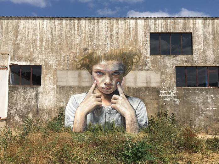 Large mural titled 'Mala hierba…' by artists Cristian Blanxer and Victor García Repo, located in Aracena, Spain. The artwork covers the weathered wall of an industrial building, featuring a young girl with a mischievous expression as she pulls down the skin below her eyes with her fingers. Rendered with realistic details and a hint of playfulness, the mural integrates with the rustic, worn surface of the building, contrasting the girl's vibrant presence against the muted, textured wall. The title 'Mala hierba…' (Bad weed…) hints at the idea of resilience or the persistent nature of wild plants.
