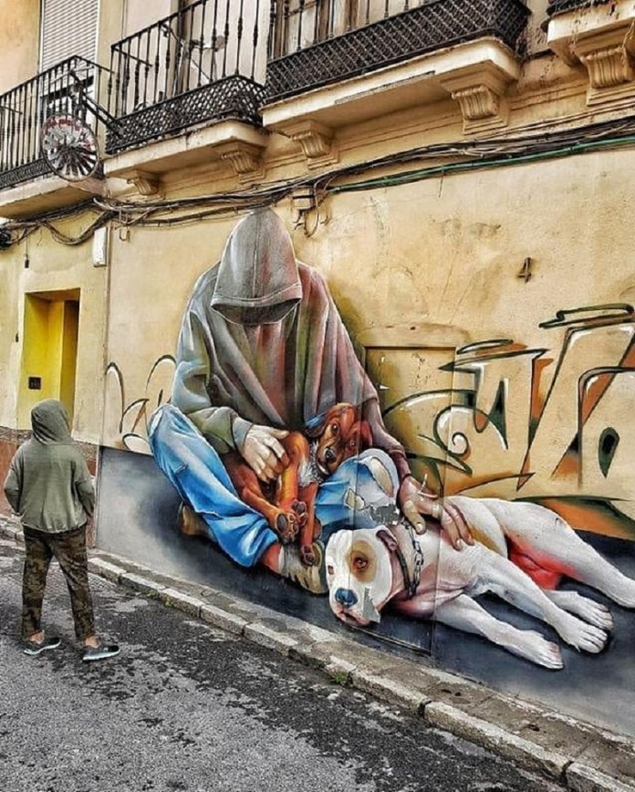 Street-level mural by Lalone showing a hooded person sitting cross-legged with two dogs, painted in realistic proportions to blend seamlessly with the urban surroundings.