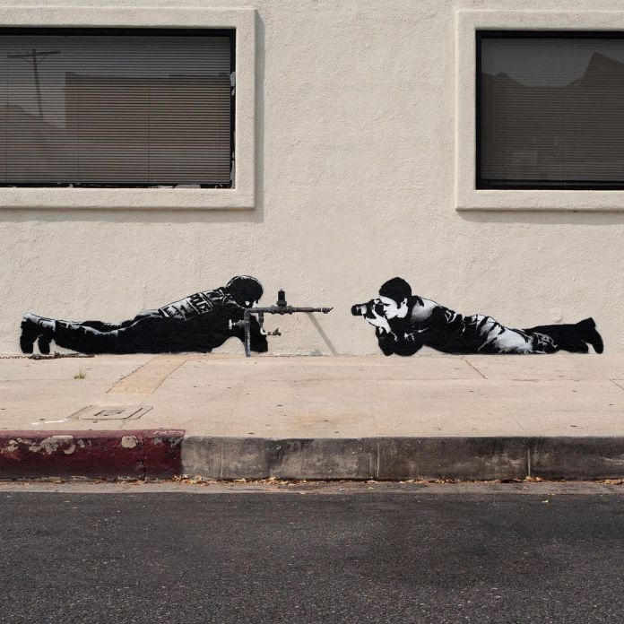 Street art stencil by Hijack showing a prone soldier aiming a gun at a prone photographer who aims a camera back, painted on a pale wall.