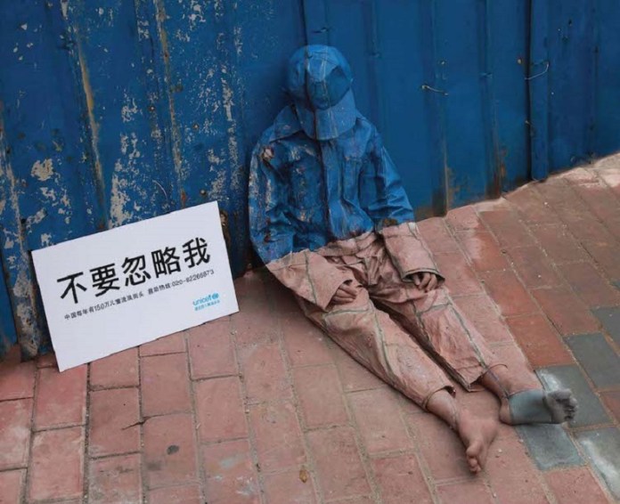 A life-like artwork from UNICEF China’s 2008 “Invisibility of Poverty” campaign shows a young boy sitting against a blue corrugated wall, painted to blend almost invisibly into his surroundings. His weathered clothing and bare feet reflect the hardships faced by homeless children. Next to him, a white sign with bold Chinese text reads “不要忽略我” (“Don’t ignore me”), accompanied by smaller text highlighting the struggles of over 1.5 million underprivileged children. The stark contrast between the boy’s camouflaged figure and the sign emphasizes the campaign’s call for awareness and action.