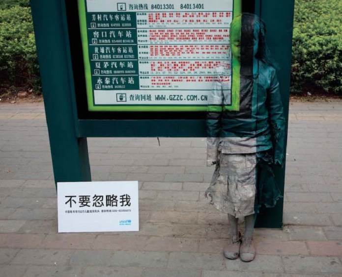 A young girl stands painted to blend into the background of a green bus stop sign as part of UNICEF China’s 2008 “Invisibility of Poverty” campaign. Her weathered clothing and solemn posture create a striking visual of how poverty renders children invisible in urban environments. Beside her, a white sign with bold Chinese text reads “不要忽略我” (“Don’t ignore me”), highlighting the struggles of over 1.5 million underprivileged children. The juxtaposition of the girl’s camouflaged figure and the visible sign emphasizes the campaign’s call for empathy and action.
