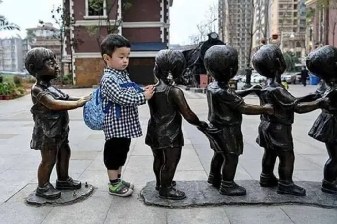 A real child with a blue backpack stands between two bronze children statues holding hands in a queue formation on a city sidewalk in China.