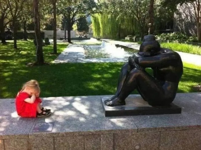 A young girl in a red dress sits with her head down next to a seated bronze statue in the same posture in a tree-lined garden with a water feature in the background.