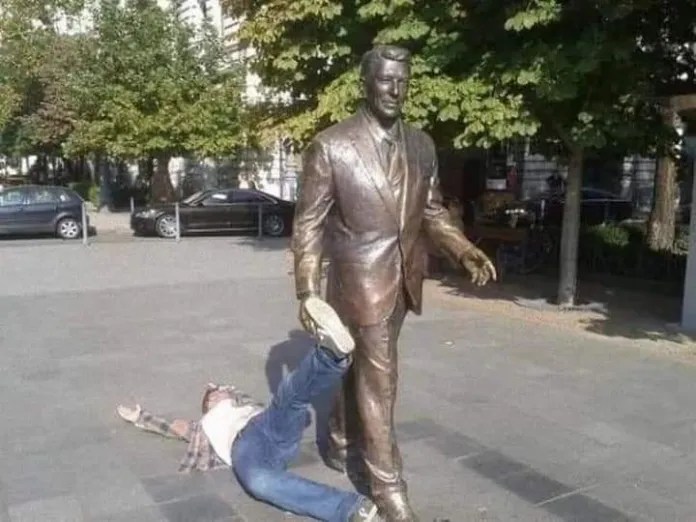 A man lies flat on his back with one leg raised and his foot placed in the hand of a walking Ronald Reagan bronze statue, creating the illusion that the statue is dragging him forward in a city square.