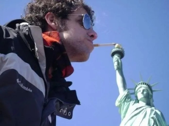 A man wearing sunglasses and a scarf poses so the flame of the Statue of Liberty lines up with a cigarette in his mouth, creating a visual illusion of it being lit.