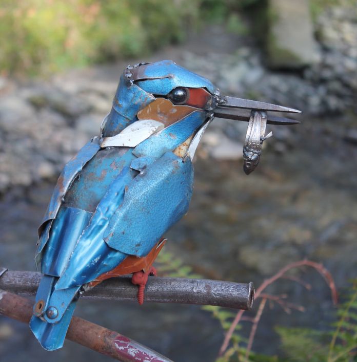 Metal sculpture of a kingfisher perched on a metal rod, created by JK Brown in the UK. The bird is crafted from blue and rust-colored scrap metal pieces and holds a small silver fish in its beak, with a rocky stream blurred in the background.