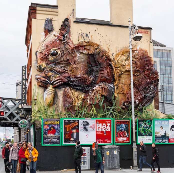Large-scale 3D mural of a red squirrel climbing a building facade, made from garbage, rusted metal, and discarded plastics, by Bordalo II in Dublin, Ireland, above a series of concert posters and urban street traffic.