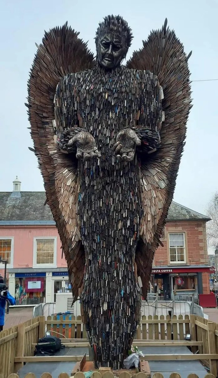 Giant angel sculpture with detailed wings and body made from thousands of real knives, standing in a fenced public square in the UK with a sorrowful face and outstretched palms, by Alfie Bradley.