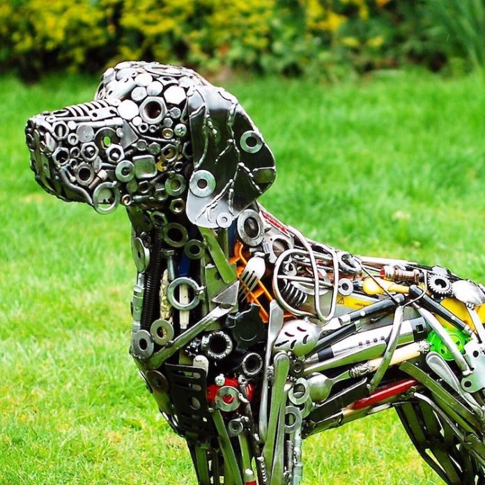 Realistic dog sculpture made entirely from recycled metal parts like screws, sockets, forks, and gears, created by Brian Mock, placed on green grass with a garden background.