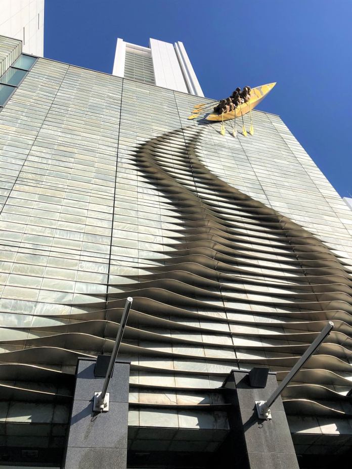 Sculpture of a yellow canoe with paddling figures appearing to climb a skyscraper in Osaka, Japan, with the wall's surface shaped into wave-like ripples for a surreal effect.