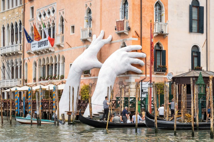 Giant white sculpture hands emerging from the water and supporting the side of a Venetian building along the Grand Canal, symbolizing environmental support.