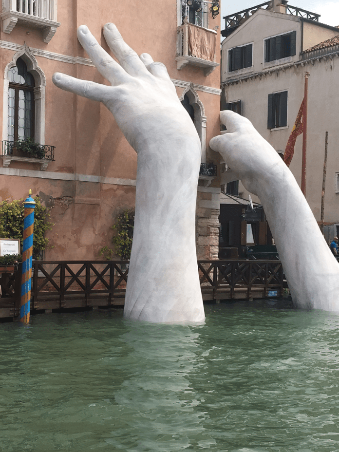 Giant white sculpture hands emerging from the water and supporting the side of a Venetian building along the Grand Canal, symbolizing environmental support.