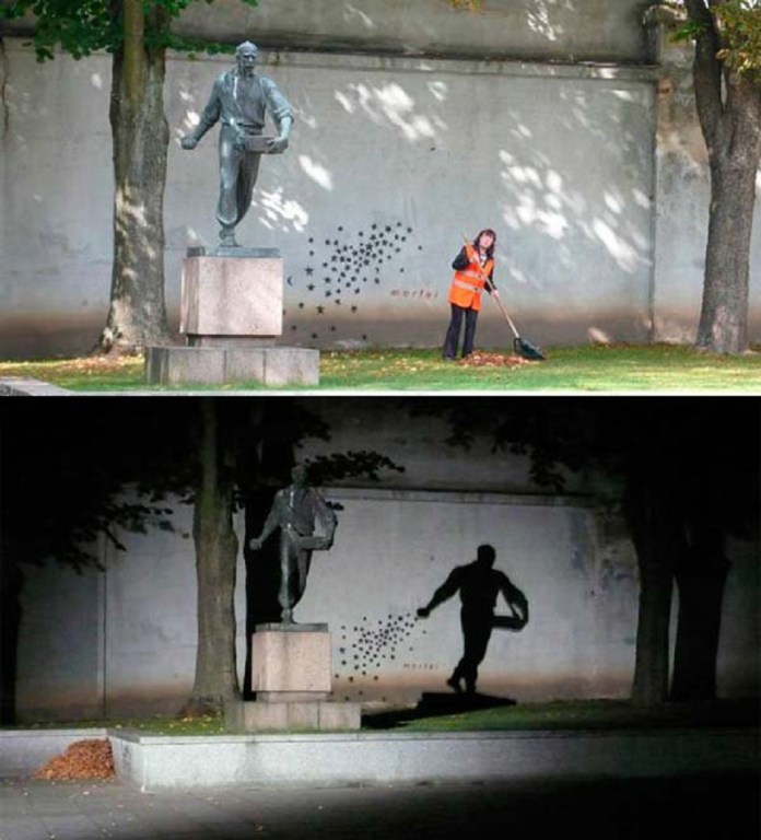 Bronze statue of a man sowing seeds in Kaunas, Lithuania, with nighttime lighting casting a shadow that transforms the gesture into spreading stars on a wall.