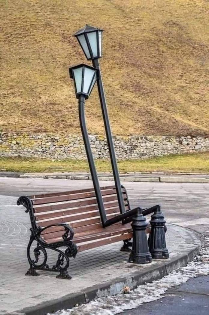Sculpture of two bent street lamps shaped like hugging figures seated on a park bench, placed along a curved sidewalk in a public space.