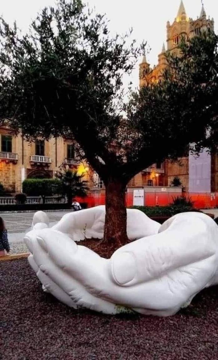 White sculpted hands cradling the base of a tree in a public square, with the surrounding buildings and cobblestone walkway in the background.