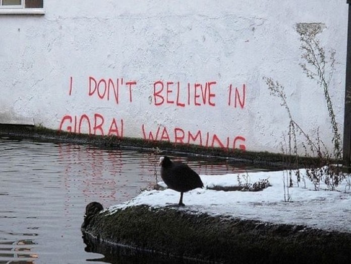 Street art by Banksy showing red text on a white wall reading “I DON'T BELIEVE IN GLOBAL WARMING,” with the lower half of the words disappearing below rising canal water in London.