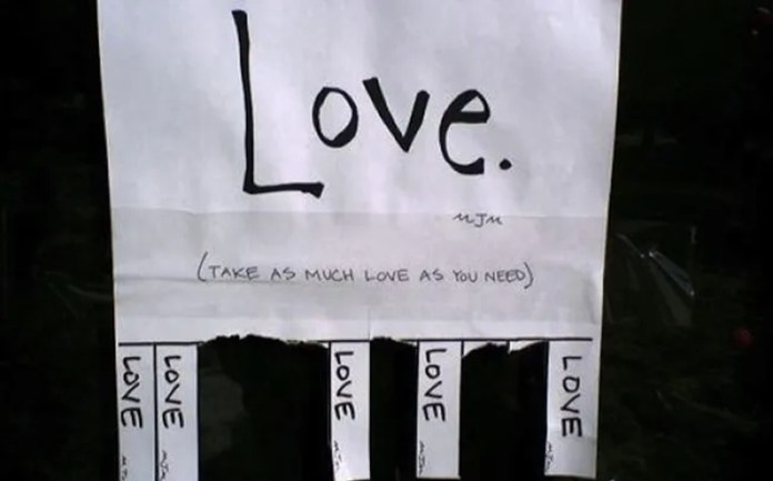 Flyer taped to a wall says “Love.” with the message “Take as much love as you need” written below, and tearable tabs labeled “LOVE.”