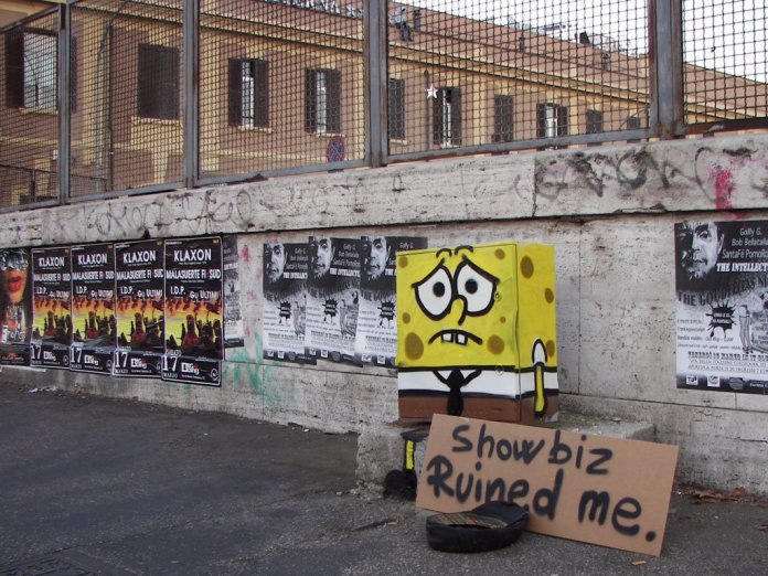 Street art sculpture of a sad SpongeBob seated beside a sign that reads “Showbiz ruined me,” placed on a Rome sidewalk against a graffiti-covered wall.