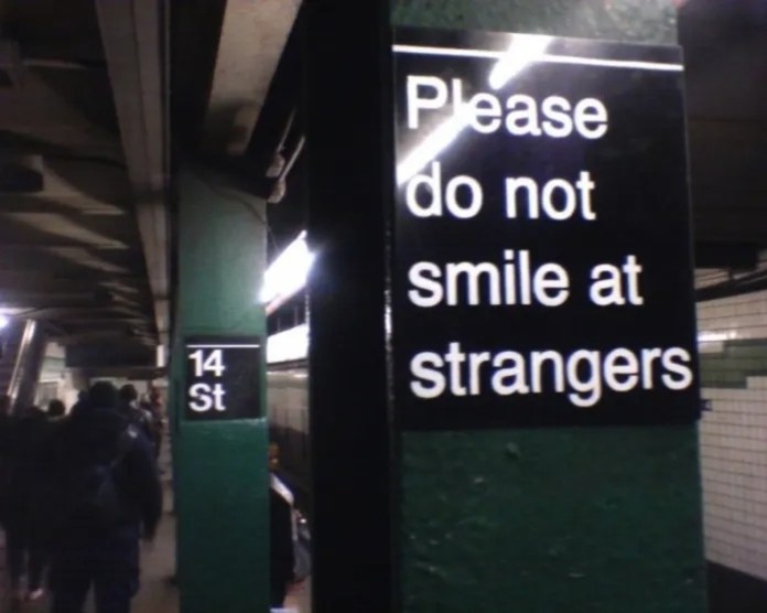 Black subway sign in New York City says “Please do not smile at strangers,” mounted to a green pillar at 14th Street station.