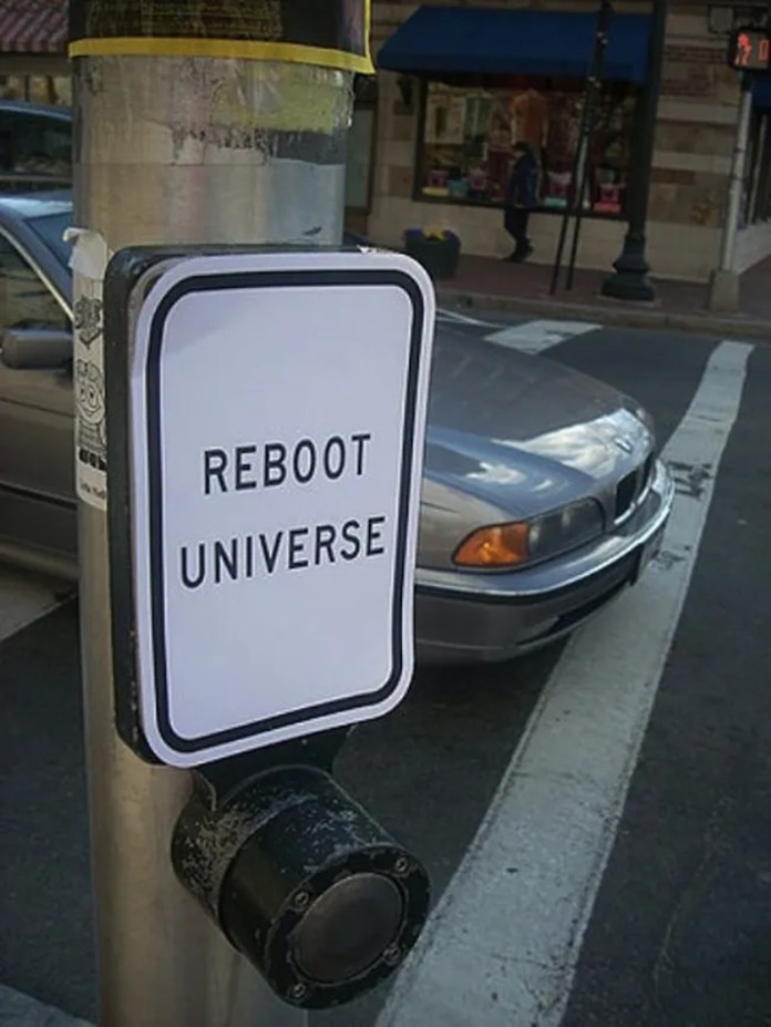 Paper sign taped over a crosswalk button reads “REBOOT UNIVERSE” in bold black letters, replacing the usual crossing instructions.