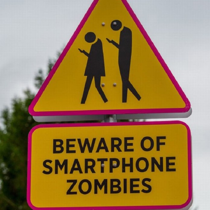 Yellow warning sign showing two human figures walking while looking at their smartphones, with bold text underneath: “BEWARE OF SMARTPHONE ZOMBIES.”