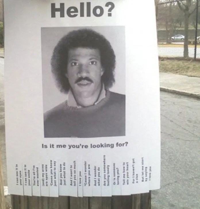 A parody flyer posted on a wooden pole featuring a black-and-white photo of Lionel Richie with the text “Hello? Is it me you're looking for?” and tear-off lyric strips referencing his famous song “Hello.”