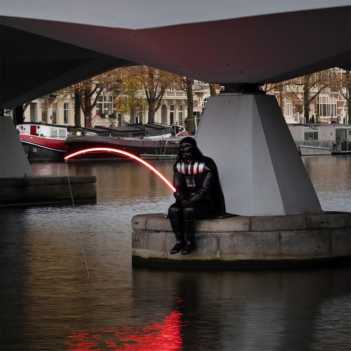 A black sculpture of Darth Vader sitting by the water under a bridge in Amsterdam, Netherlands, holding a red lightsaber that curves like a fishing rod, with its glow reflecting in the canal below.