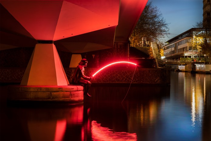 Street art sculpture Darth Fisher by Frankey, located at De Torontobrug (Bridge 350) in Amsterdam, Netherlands. The piece features Darth Vader sitting on a concrete ledge beneath a bridge, casually fishing with a glowing red lightsaber instead of a fishing rod. The red glow reflects on the water, adding a striking sci-fi touch to the urban setting.