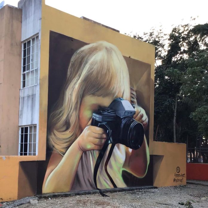 Mural on a building in Playa del Carmen showing a young blonde girl holding a DSLR camera in front of her face, painted in hyper-realistic style by Hebs Art.