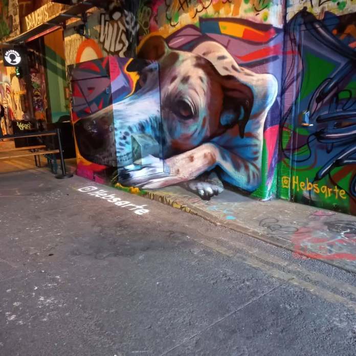 Photorealistic 3D mural of a dog lying across a wall and electrical box in Leake Street Tunnel, London. The realistic posture and shadows make it appear life-sized and real.