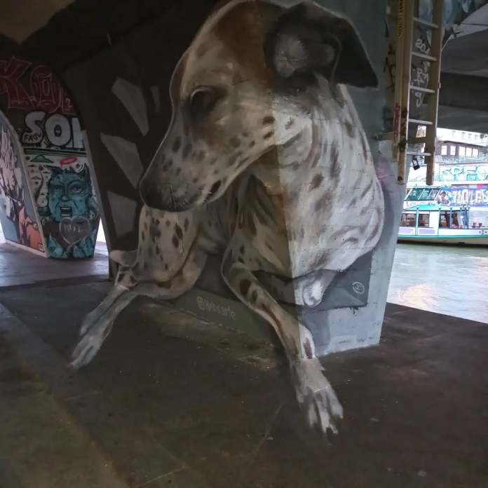 A lounging dog painted in realistic 3D perspective by Hebs Art on a concrete pillar by the river in Vienna, Austria. The dog’s form wraps around the shape of the pillar with believable lighting and fur texture.