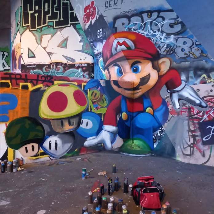 Street art mural of Super Mario jumping, surrounded by mushrooms and power-ups, painted under a bridge in Stadlau, Vienna. The piece is layered over older graffiti and integrates game icons.